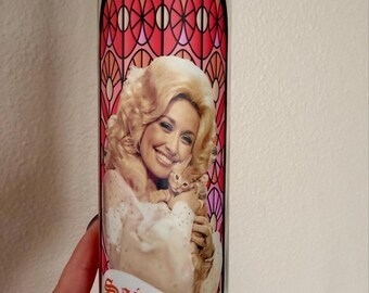 Puede incluir: Una vela de oración cilíndrica de vidrio rellena de cera rosa. La etiqueta presenta el retrato de una mujer rubia sosteniendo un gatito pequeño, sobre un patrón de vidrieras rojas y rosas. El texto "Saint Dolly" está impreso en rojo.