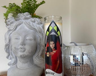 Könnte beinhalten: Eine Glaskerze mit einem Etikett mit einem Porträt einer Frau in einem roten Kleid, mit dem Text "Saint Lydia". Die Kerze steht neben einem grauen Steinbüsten-Pflanzgefäß mit grünen Farnen und einem silbernen Discokugel-Topf.
