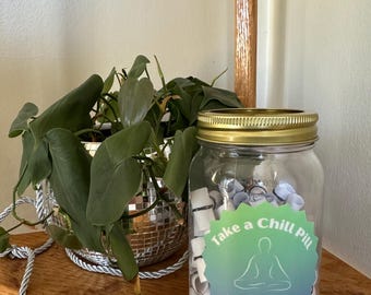 Könnte beinhalten: Ein Glasgefäß gefüllt mit aufgerollten Papierstücken mit schwarzer Schrift. Das Gefäß hat einen goldenen Deckel und einen grün-blauen Aufkleber mit der Aufschrift "Take a Chill Pill" und einer Zeichnung einer Person in Meditationshaltung. Der Aufkleber sagt auch "A jar of affirmations for when you need to just Take a Chill Pill."