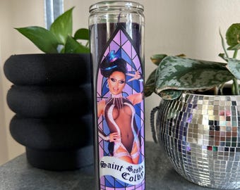 Könnte beinhalten: Eine Glaskerze mit einem violetten und blauen Buntglasdesign, das ein Porträt einer Person mit dunklen Haaren und einem schwarzen Outfit zeigt. Der Text "Saint Sasha Colby" ist auf die Kerze gedruckt.