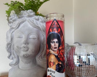 Puede incluir: Una vela roja en un tarro de cristal transparente, con una imagen de una persona con cabello oscuro y el texto "Saint Frank-N-Furter". También se ve una estatua gris de una cabeza de mujer y una bola de discoteca espejada.