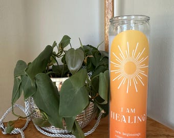 Könnte beinhalten: Eine weiße Kerze mit einem gelben und orangefarbenen Etikett, auf dem "I AM HEALING" steht, darunter der Text "new beginnings, relieve anxieties, optimize power". Die Kerze steht auf einem Holzregal mit einer grünen Pflanze in einem silbernen Topf.