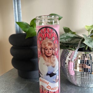 Puede incluir: Vela de vidrio rosa con un dise&ntilde;o de vitral que presenta un retrato de Dolly Parton. La vela tiene el texto "Saint Dolly" impreso en la parte delantera.