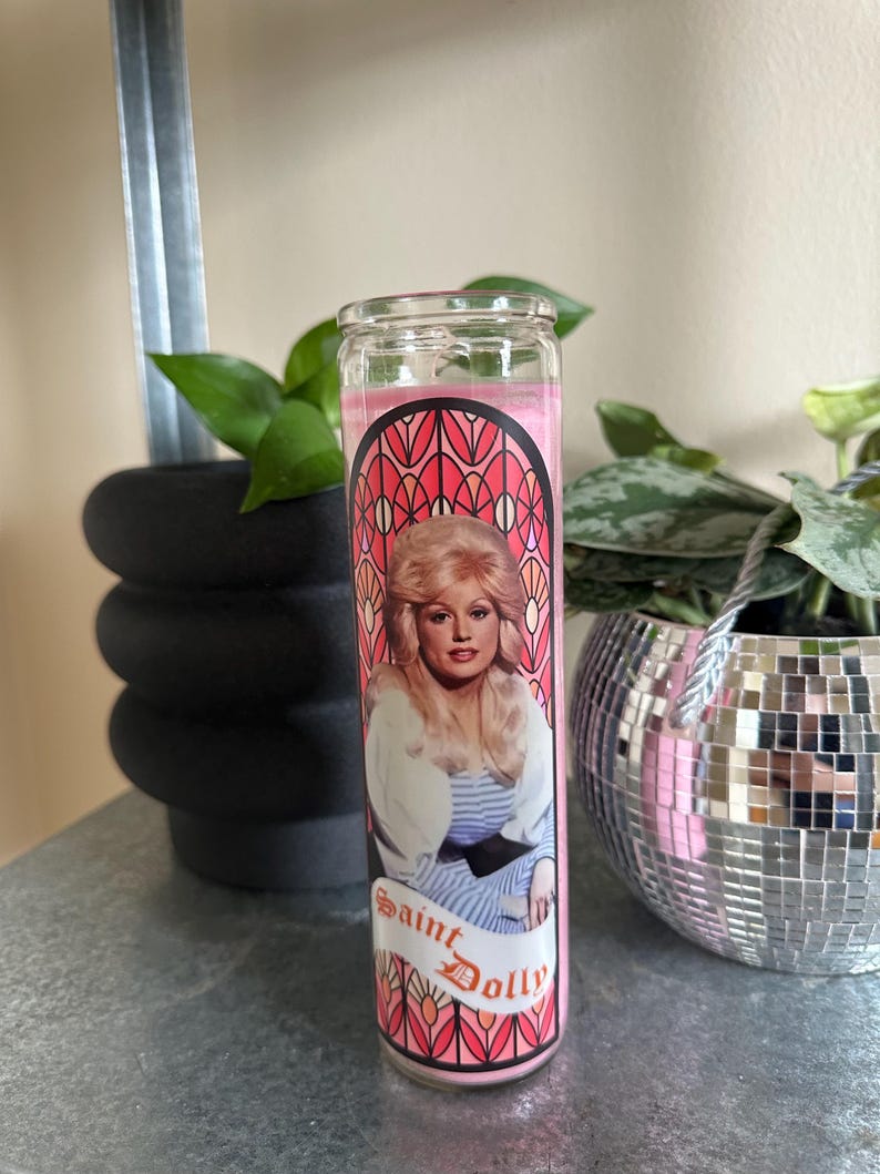 Puede incluir: Vela de vidrio rosa con un dise&ntilde;o de vitral que presenta un retrato de Dolly Parton. La vela tiene el texto "Saint Dolly" impreso en la parte delantera.