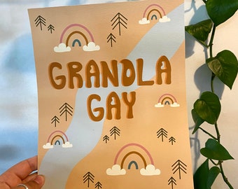 Könnte beinhalten: Ein Pfirsichfarbener Druck mit dem Text "Grandla Gay" in braunen Buchstaben. Der Druck zeigt einen stilisierten blauen Fluss mit weißen Wolken und Regenbögen in Rosa, Braun und Blau. Es gibt auch kleine braune Bäume auf dem gesamten Druck.