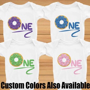 May include: Four white baby onesies with a donut graphic and the word "ONE" in different colors. The donuts are purple, blue, green, and pink. The text is in purple, blue, green, and pink to match the donuts. The text "Custom Colors Also Available" is at the bottom of the image.