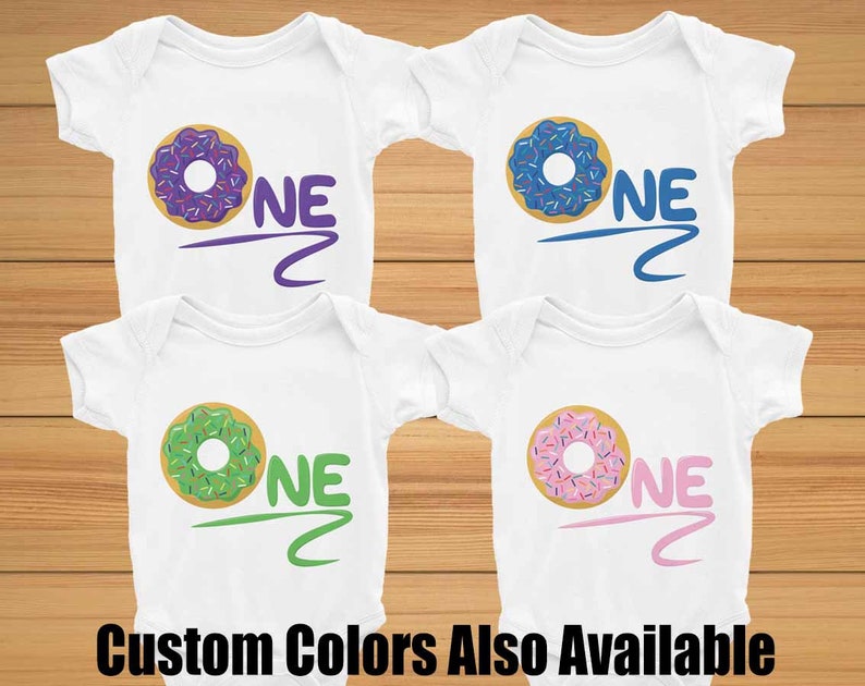 May include: Four white baby onesies with a donut graphic and the word "ONE" in different colors. The donuts are purple, blue, green, and pink. The text is in purple, blue, green, and pink to match the donuts. The text "Custom Colors Also Available" is at the bottom of the image.