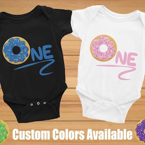 May include: Two baby onesies with a donut graphic and the word "ONE" in a colorful font. The onesies are black and white. The donut on the black onesie is blue with colorful sprinkles. The donut on the white onesie is pink with colorful sprinkles. Custom colors are available.