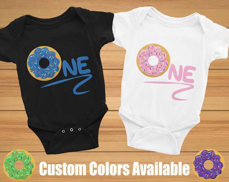 May include: Two baby onesies with a donut graphic and the word "ONE" in a colorful font. The onesies are black and white. The donut on the black onesie is blue with colorful sprinkles. The donut on the white onesie is pink with colorful sprinkles. Custom colors are available.