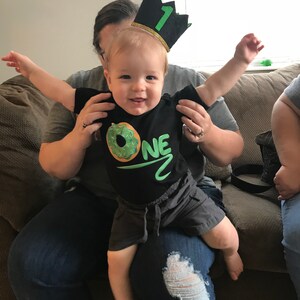 May include: A baby wearing a black shirt with a green donut and the word "ONE" on it. The baby is wearing a black and green crown with the number "1" on it. The baby is being held by a person wearing blue jeans.