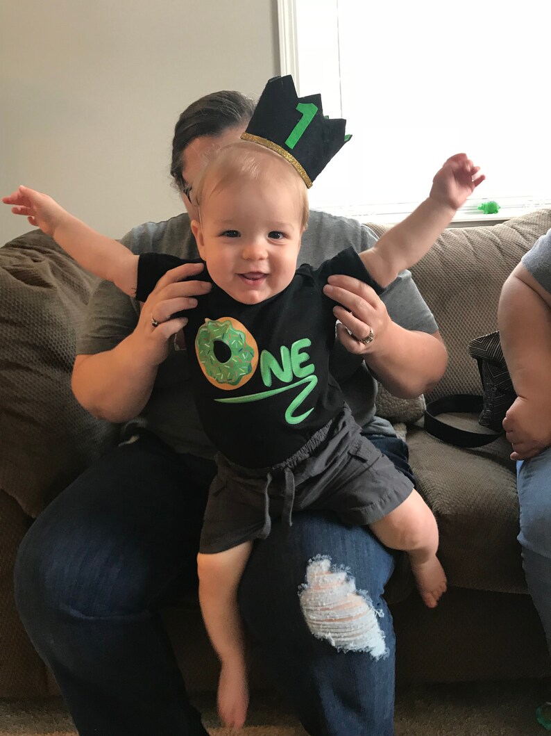 May include: A baby wearing a black shirt with a green donut and the word "ONE" on it. The baby is wearing a black and green crown with the number "1" on it. The baby is being held by a person wearing blue jeans.