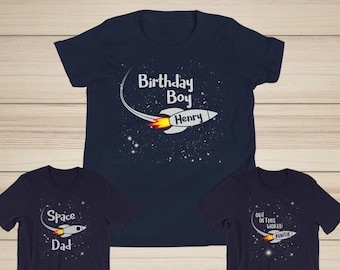May include: Three navy blue t-shirts with a space theme. The first shirt says "Birthday Boy Henry" with a rocket ship. The second shirt says "Space Dad" with a rocket ship. The third shirt says "Out of this world Auntie" with a rocket ship.