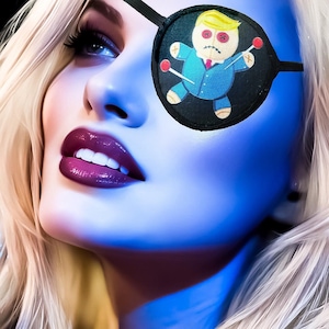 May include: A woman with blonde hair and dark purple lipstick wears a black eye patch with a cartoon voodoo doll design. The doll is wearing a blue suit and has red pins.