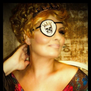 May include: A woman wearing a gold crown and a skull eye patch, with a floral and lace top.