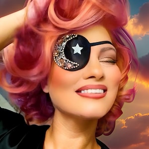 May include: A person with pink and purple hair wearing a black eye patch decorated with a crescent moon and star design, embellished with rhinestones. The person is smiling, with a sunset sky in the background.