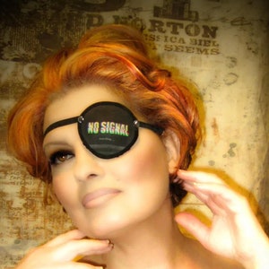 May include: A woman with red hair is wearing a black eye patch that says "NO SIGNAL". She is wearing a black off-the-shoulder top.