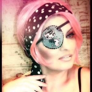 May include: A woman with pink hair is wearing a black and white polka dot headband and a black eye patch with a cartoon cat design. She is wearing a black tank top.