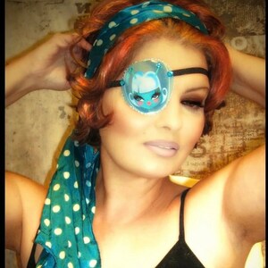 May include: A woman with red hair is wearing a blue and white polka dot headscarf and a black tank top. She is wearing a blue and white eye patch with a cartoon character on it.