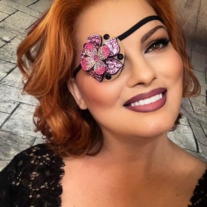 May include: A woman with red hair is wearing a black eye patch with a pink and white flower design. The flower is covered in rhinestones.