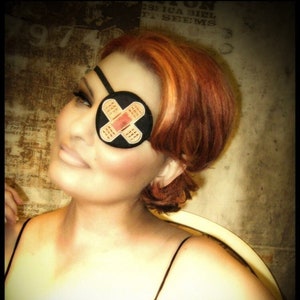 May include: A woman with red hair is wearing a black eye patch with a white bandage on it. She is wearing a black spaghetti strap top.