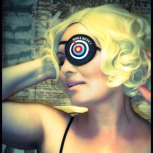 May include: A woman with blonde hair is wearing a black dress with a lace overlay and a black eye patch that says "Bullseye!" The eye patch has a red, white, and blue target design.