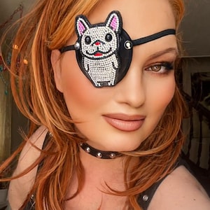 May include: Black leather eye patch with a beaded French bulldog design. The eye patch is worn with a black studded choker and a black top with harness straps. The person has red hair and is wearing makeup.