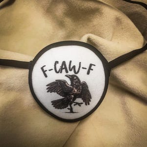 May include: An eye patch with a black border and elastic strap. The white center features a black raven illustration and the text "F-CAW-F". The eye patch is designed to cover one eye.