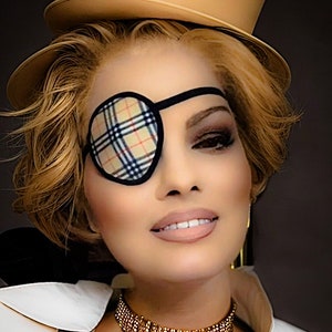 May include: A woman with blonde hair is wearing a brown top hat, a black eye patch with a plaid pattern, a gold choker necklace, and a white collared shirt.