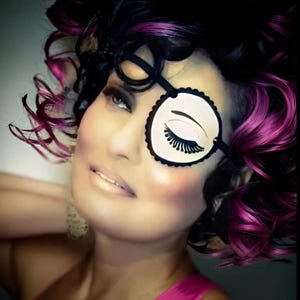 May include: A round white eye patch featuring a black graphic illustration of a closed eye with long eyelashes. The patch is secured with a black strap and worn over one eye, complemented by dark hair with vibrant magenta highlights.