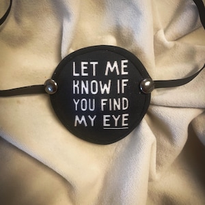 May include: A circular black eye patch featuring white embroidered text that reads, LET ME KNOW IF YOU FIND MY EYE. The patch is constructed with a black fabric border, two metallic silver rivets on the sides, and a thin black elastic strap.