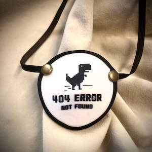 May include: White eye patch with a black border and elastic strap. The patch features a pixelated black dinosaur graphic and the text "404 ERROR NOT FOUND". Fastened with two brass-colored rivets.