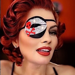 May include: A woman with red hair is wearing a black eye patch with a red skull and crossbones design and rhinestones. The eye patch is covering her right eye.