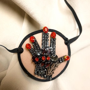 May include: A circular eye patch featuring a beige fabric base with a hand-shaped appliqué. The hand is embellished with dark grey crystals, beads, and vibrant red oval stones at the fingertips and palm, framed by a black border with thin straps.