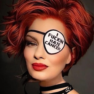 May include: A white circular eye patch with a black border and the black text I FUCKIN HATE CANCER. The patch is secured with a thin black elastic strap that wraps around the head, positioned over the left eye area.