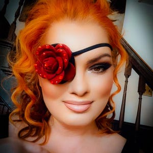 May include: An eye patch featuring a large, deep red fabric rose adorned with small, sparkling red crystals. The rose is attached to a thin, black elastic strap designed to secure the patch comfortably over one eye for a secure fit.
