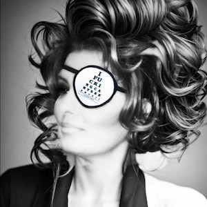May include: A black and white eye patch featuring a printed eye chart design with the text I FUCKING CANT SEE SHIT. The patch has a circular white center with black lettering and a thick black border, secured by a thin black elastic strap.