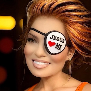 May include: A woman with red hair is wearing a black eye patch with a white circle and the text "JESUS LOVE ME" and a red heart. She is smiling and wearing a bright orange tank top.