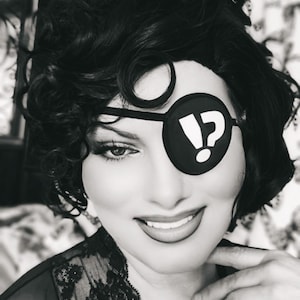 May include: Black and white portrait of a person wearing a black eye patch with a white question mark and exclamation point. The person has dark hair and is smiling, wearing a black lace top.