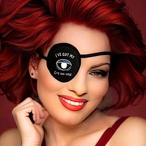 May include: A woman with bright red hair is wearing a black eye patch with the text "I've got my eye on you".