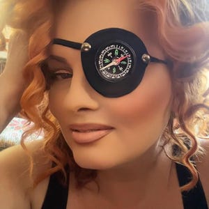 May include: A black eye patch with a compass design is worn over one eye. The compass features a white face with directional markings. The person has red curly hair and is wearing a black top.