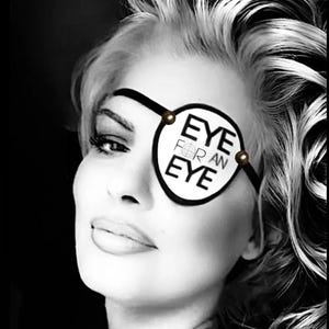 May include: A black and white eye patch featuring the text EYE FOR AN EYE with a crosshair symbol on the letter O. The patch is circular with a black border and two small brown beads attached to the black elastic strap on each side.