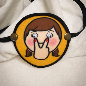 May include: A circular eye patch featuring a yellow background with a black border and a black elastic strap. The center displays an embroidered illustration of a face with brown hair, pink cheeks, and two googly eyes held by a hand.