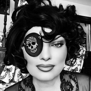 May include: Black and white image of a person wearing a black eye patch with a skull design. The eye patch is round and has a silver trim. The person has dark hair and is wearing a lace top.