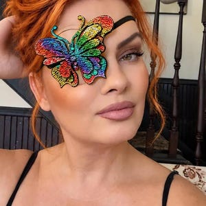 May include: An eye patch featuring a large, vibrant butterfly design covered in multicolored rhinestones. The butterfly displays a spectrum of colors including red, orange, yellow, green, blue, and purple. It is attached to a thin black elastic strap.