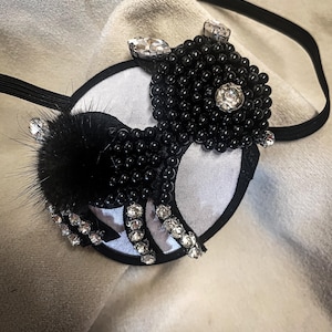 May include: A black and white eye mask with a beaded design and rhinestone accents. The mask features a black fur accent and is attached to a black elastic band. The design is centered on a white and black circular base.