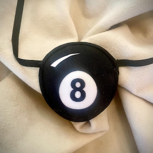 May include: Black eye patch featuring a white number 8 and a white curved stripe, resembling a billiard ball. The eye patch has a black elastic strap for securing it around the head. The eye patch is designed to cover one eye.