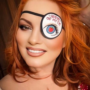 May include: A person with vibrant red hair and a decorative eye patch. The eye patch is white with a blue eye and a red apple design, and the text "apple of my eye". The person is wearing a red dress with white polka dots.