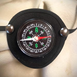 May include: A black circular eye patch featuring a centered magnetic compass with a clear glass face. The compass dial displays cardinal directions in green and white text, with a red and white needle. Two silver-toned metal rivets secure the elastic strap.
