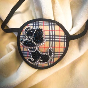 May include: An eye patch with a black elastic band. The patch features a tan, red, and black plaid pattern with a black and crystal-studded bear design. The patch is circular with a black border.