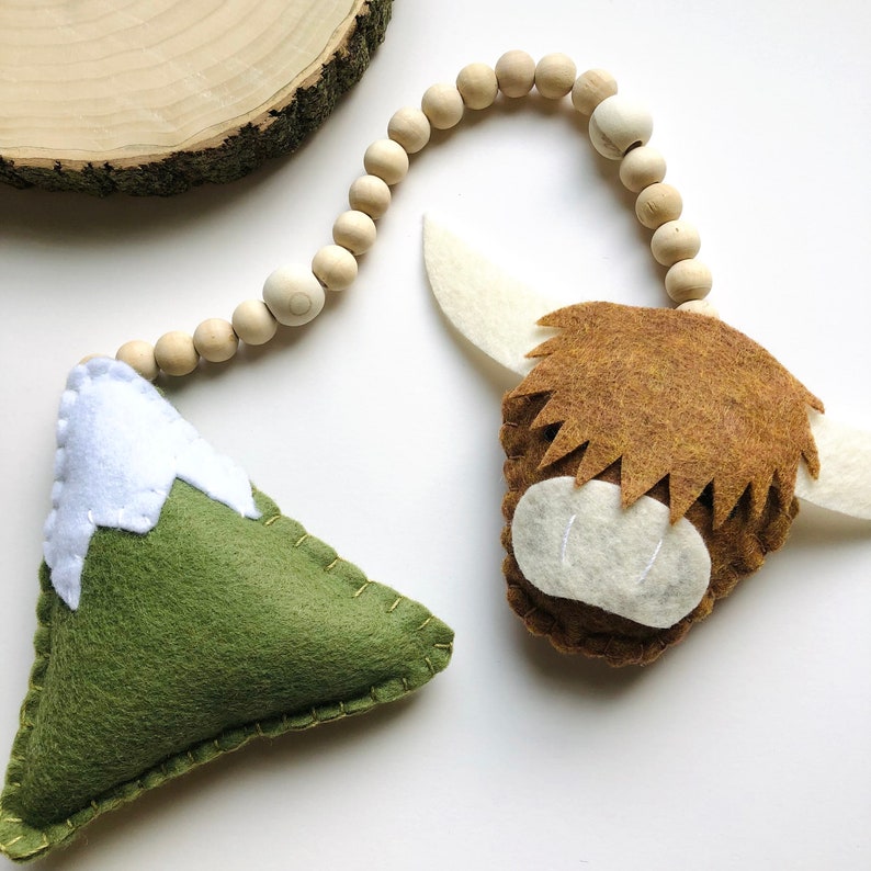 May include: A felt mountain with a white snowy peak and a brown highland cow head with white facial features are strung together with a wooden bead garland.